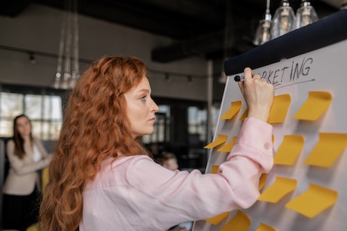 A woman writing on a whiteboard covered in yellow sticky notes.